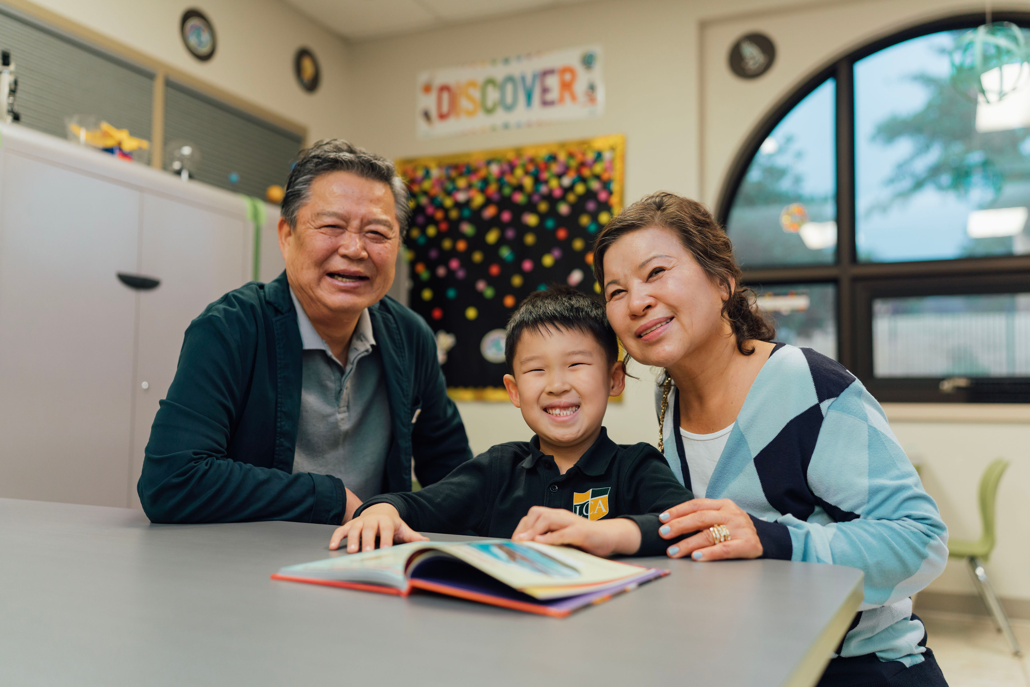 Grandparents sitting with their grandchild at a classroom table, looking at a book together.