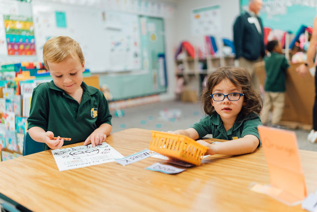 Two students working together at a classroom table while a guest visits.