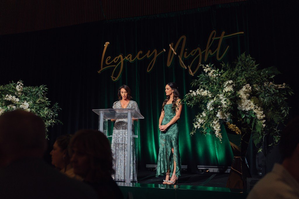 Hosts on stage beneath Legacy Night gold script at the gala