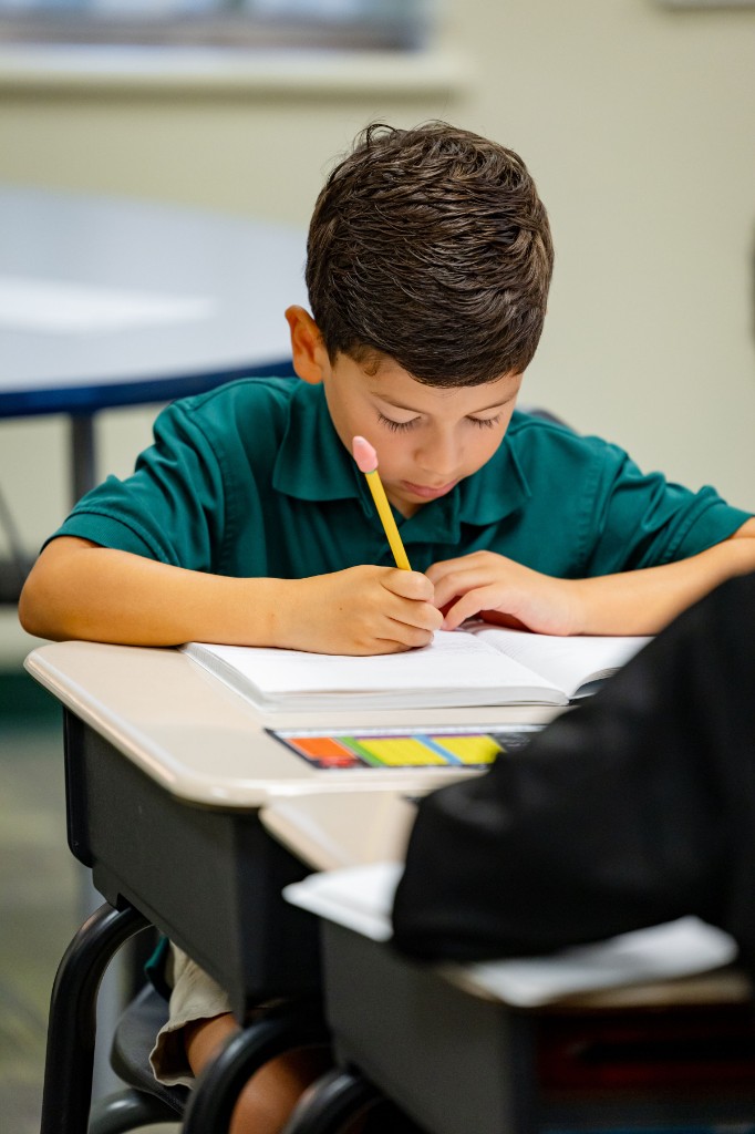A young Legacy Christian Academy student writing in a notebook at his classroom desk